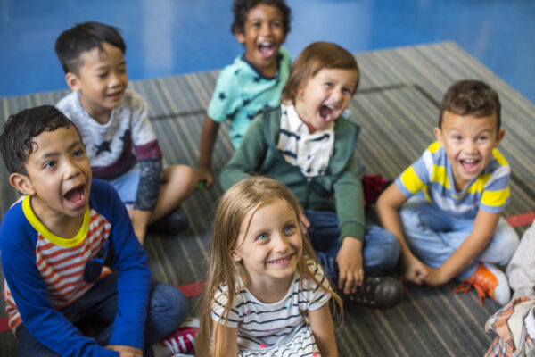 A diverse group of young children smiles and laughs on a striped rug, with a blue backdrop, embodying the principles taught in the BCFCCA: 20 Hour Responsible Adult: Professional Development for Child Care Providers Level 1 program.
